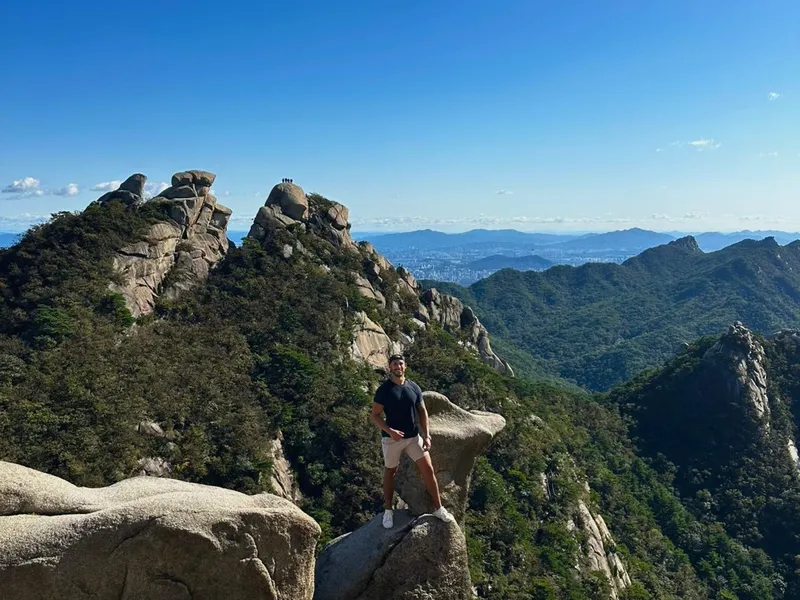Bruno standing on a granite summit in Bukhansan with forested peaks and the Seoul skyline in the distance